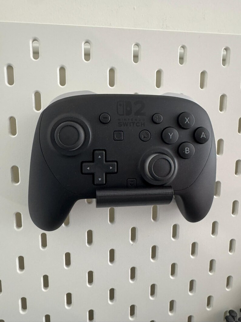 K&ouml;nnte beinhalten: Ein schwarzer Nintendo Switch Pro Controller mit grauen Akzenten ist an einer wei&szlig;en Lochwand befestigt. Der Controller verf&uuml;gt &uuml;ber ein Steuerkreuz, Tasten mit den Bezeichnungen X, Y, A und B sowie zwei Analog-Sticks. Das Nintendo Switch-Logo ist sichtbar.