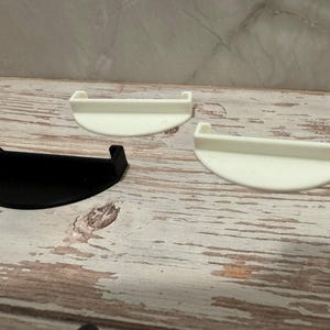 May include: Three plastic, semi-circular shelves. One is black, and the other two are white. Each shelf has a raised edge on either side. The shelves are displayed on a weathered, light-colored wooden surface.