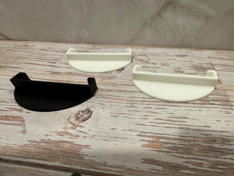 May include: Three plastic, semi-circular shelves. One is black, and the other two are white. Each shelf has a raised edge on either side. The shelves are displayed on a weathered, light-colored wooden surface.