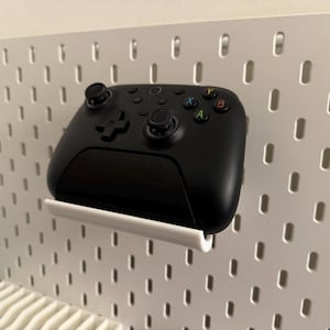 Puede incluir: Un mando de videojuegos negro está montado en una pared perforada blanca con un soporte blanco. El mando tiene botones de colores y dos joysticks. La pared perforada tiene agujeros espaciados.