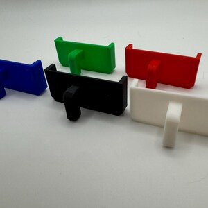 May include: Five 3D printed phone stands in different colours: blue, green, black, white, and red. Each stand has a small lip on the front to hold a phone.