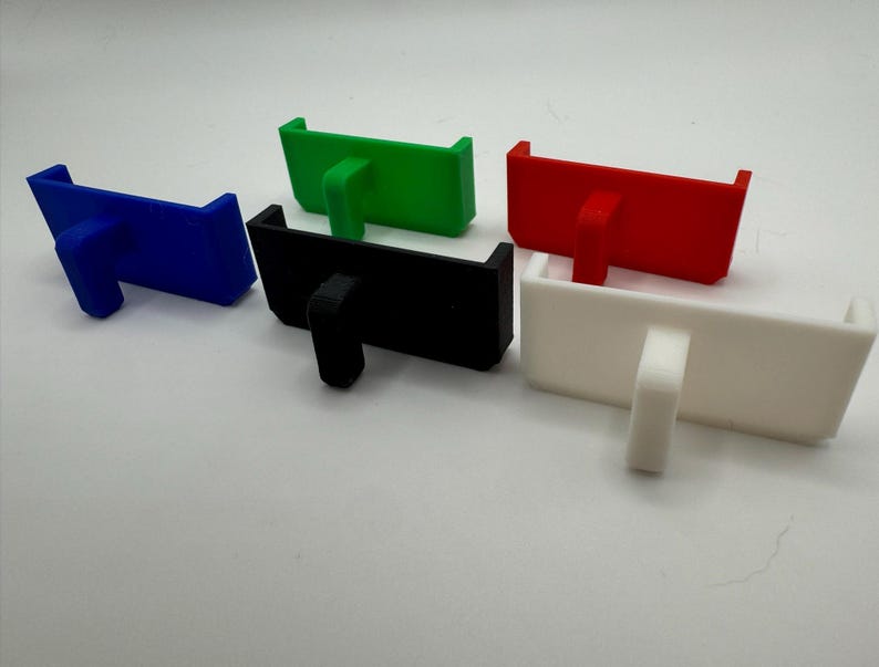 May include: Five 3D printed phone stands in different colours: blue, green, black, white, and red. Each stand has a small lip on the front to hold a phone.