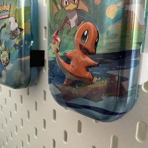 K&ouml;nnte beinhalten: Zwei blaue Metall-Wasserflaschen mit Pok&eacute;mon-Grafiken. Eine zeigt Charmander, die andere andere Pok&eacute;mon-Charaktere. Die Flaschen h&auml;ngen an einer wei&szlig;en Lochwand. Die Flaschen haben schwarze und wei&szlig;e Clips.