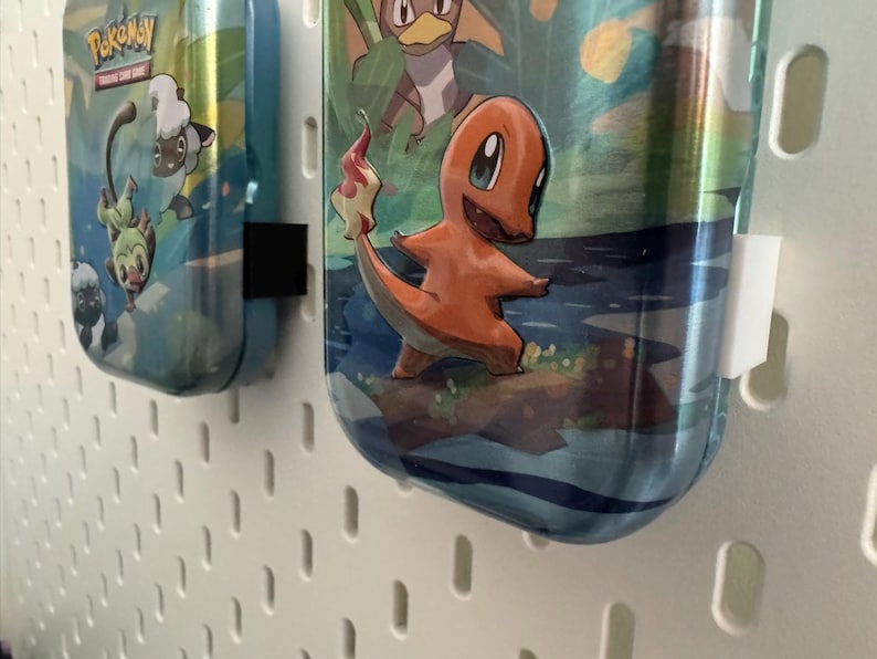 K&ouml;nnte beinhalten: Zwei blaue Metall-Wasserflaschen mit Pok&eacute;mon-Grafiken. Eine zeigt Charmander, die andere andere Pok&eacute;mon-Charaktere. Die Flaschen h&auml;ngen an einer wei&szlig;en Lochwand. Die Flaschen haben schwarze und wei&szlig;e Clips.