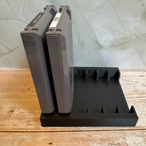 May include: A black, 3D-printed video game cartridge holder with slots for multiple cartridges. Two gray game cartridges are inserted into the holder. The holder is on a wooden surface.