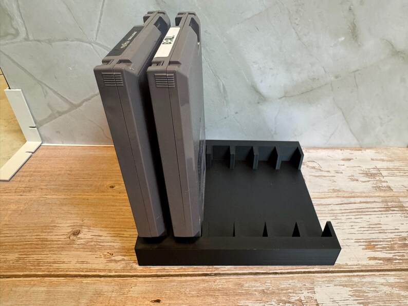 May include: A black, 3D-printed video game cartridge holder with slots for multiple cartridges. Two gray game cartridges are inserted into the holder. The holder is on a wooden surface.