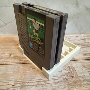 May include: A white 3D-printed game cartridge holder with three dark gray Nintendo Entertainment System game cartridges. The front cartridge displays the game "Tennis" with a player graphic. The holder is on a light wood surface.