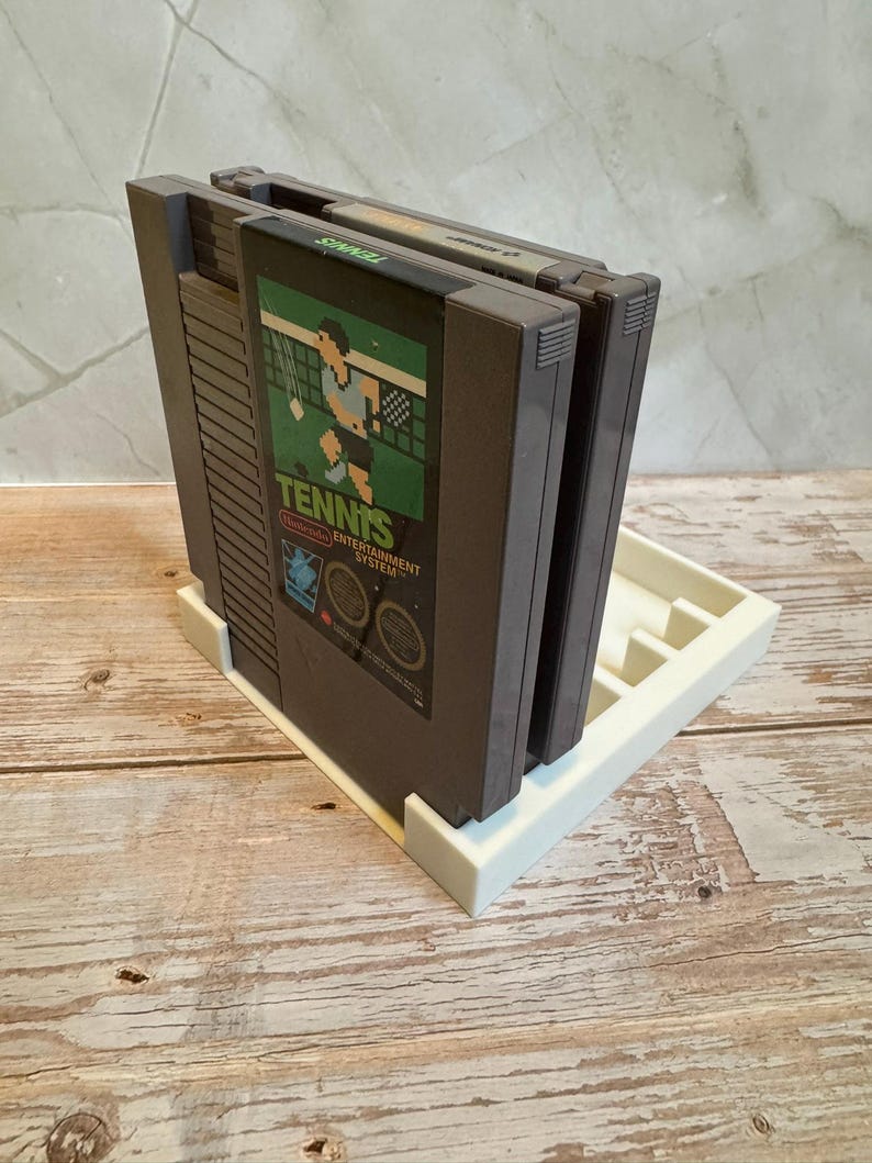May include: A white 3D-printed game cartridge holder with three dark gray Nintendo Entertainment System game cartridges. The front cartridge displays the game "Tennis" with a player graphic. The holder is on a light wood surface.