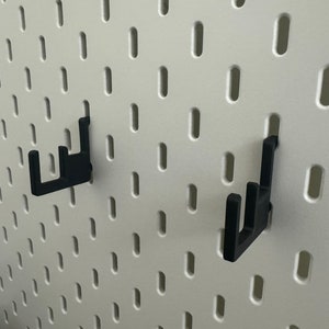 May include: Two black plastic hooks mounted on a white pegboard wall.