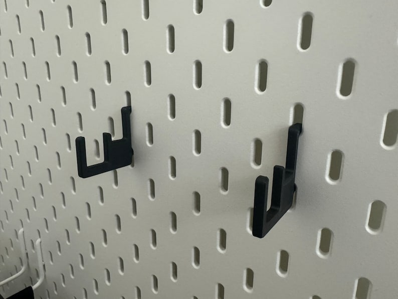May include: Two black plastic hooks mounted on a white pegboard wall.