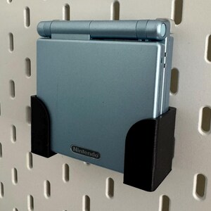 Pu&ograve; includere: Una console per videogiochi portatile Game Boy Advance SP blu con un supporto da parete nero. La console ha il logo Nintendo sul davanti.