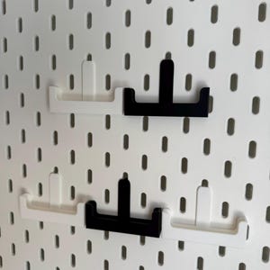Puede incluir: Cuatro ganchos para panel perforado en blanco y negro se muestran sobre un fondo de panel perforado blanco. Los ganchos est&aacute;n dise&ntilde;ados para encajar en los agujeros del panel perforado. Dos ganchos son blancos y dos son negros. Los ganchos son de material s&oacute;lido.