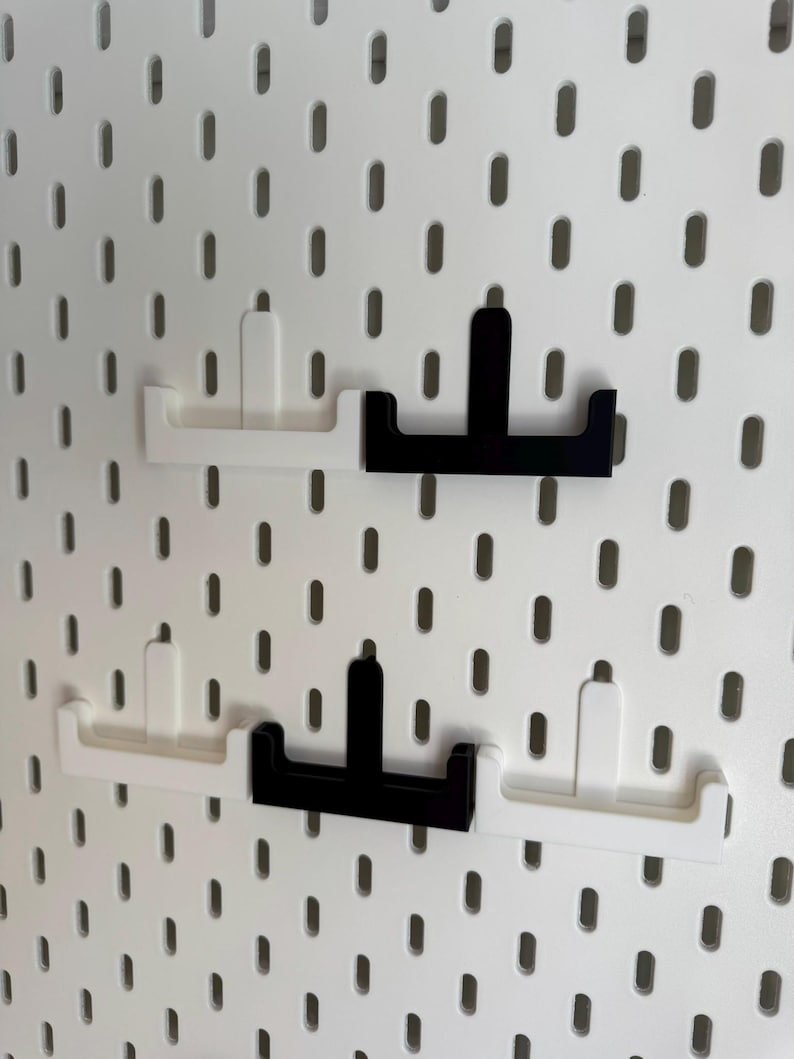 Puede incluir: Cuatro ganchos para panel perforado en blanco y negro se muestran sobre un fondo de panel perforado blanco. Los ganchos est&aacute;n dise&ntilde;ados para encajar en los agujeros del panel perforado. Dos ganchos son blancos y dos son negros. Los ganchos son de material s&oacute;lido.