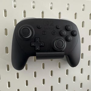K&ouml;nnte beinhalten: Ein schwarzer Nintendo Switch Pro Controller ist an einer wei&szlig;en Lochwand befestigt. Der Controller hat zwei Analog-Sticks, ein Steuerkreuz und Aktionstasten. Das Nintendo Switch-Logo ist sichtbar.