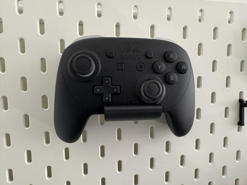 K&ouml;nnte beinhalten: Ein schwarzer Nintendo Switch Pro Controller ist an einer wei&szlig;en Lochwand befestigt. Der Controller hat zwei Analog-Sticks, ein Steuerkreuz und Aktionstasten. Das Nintendo Switch-Logo ist sichtbar.