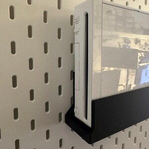 Peut inclure: Une console de jeu blanche mont&eacute;e sur un panneau perfor&eacute; blanc avec un support noir. La console porte le mot "ATI". Le panneau perfor&eacute; pr&eacute;sente des trous ovales espac&eacute;s uniform&eacute;ment.