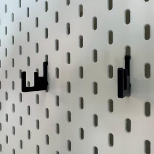May include: Two black pegboard hooks are attached to a white pegboard. The hooks are made of a solid material and are designed to hold items. The pegboard has evenly spaced oval holes.