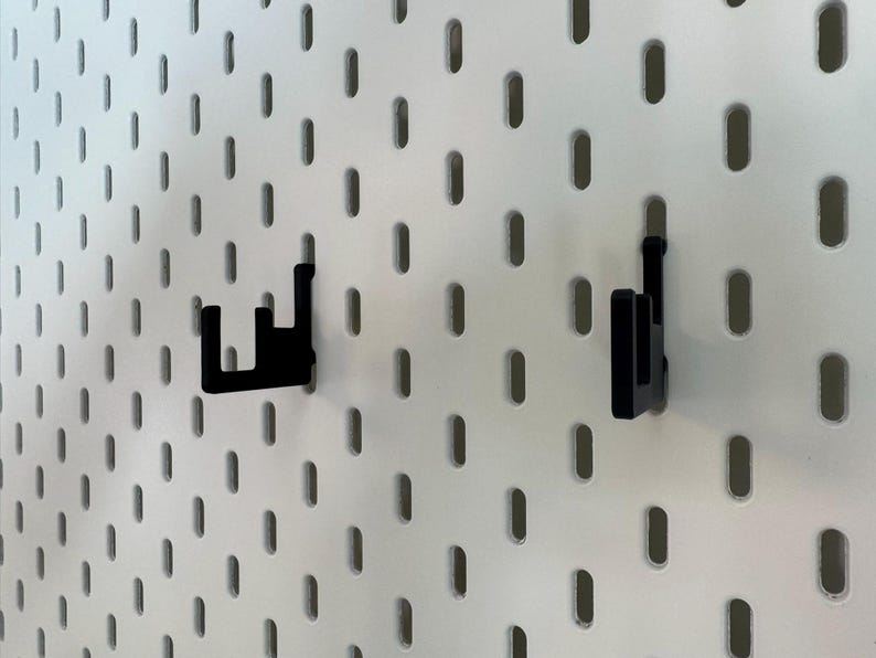 May include: Two black pegboard hooks are attached to a white pegboard. The hooks are made of a solid material and are designed to hold items. The pegboard has evenly spaced oval holes.