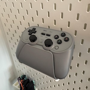 Peut inclure: Une manette de jeu Pro 3 sans fil grise avec des boutons noirs et un pavé directionnel, montée sur un panneau perforé blanc. La manette a un design élégant, idéale pour les jeux.
