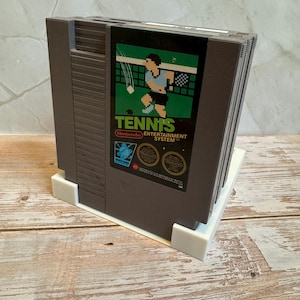 May include: A stack of Nintendo Entertainment System (NES) game cartridges. The front cartridge features the game "Tennis" with a pixelated tennis player graphic. The cartridges are dark gray and rest on a white plastic stand.