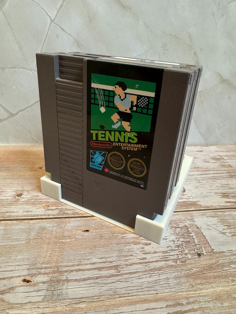 May include: A stack of Nintendo Entertainment System (NES) game cartridges. The front cartridge features the game "Tennis" with a pixelated tennis player graphic. The cartridges are dark gray and rest on a white plastic stand.