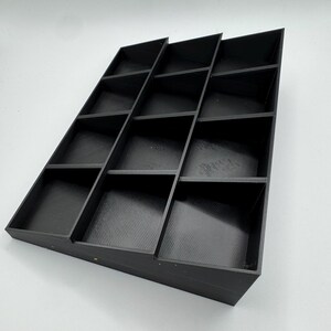 Peut inclure: Un organisateur noir inclin&eacute; avec plusieurs compartiments rectangulaires. L'organisateur est fait d'un mat&eacute;riau solide et est con&ccedil;u pour contenir de petits objets. Les compartiments sont dispos&eacute;s en gradins.