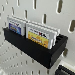 Op de afbeelding: Een zwarte, rechthoekige plank ontworpen om Nintendo 3DS-gamecartridges in op te bergen. De plank is gemonteerd op een wit pegboard. Verschillende gamecartridges zijn zichtbaar, waaronder "Detective Pikachu" en "Pokemon X".