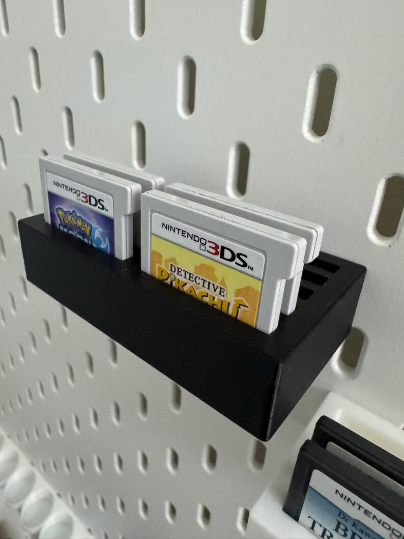Op de afbeelding: Een zwarte, rechthoekige plank ontworpen om Nintendo 3DS-gamecartridges in op te bergen. De plank is gemonteerd op een wit pegboard. Verschillende gamecartridges zijn zichtbaar, waaronder "Detective Pikachu" en "Pokemon X".