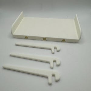May include: White plastic shelf with three white plastic brackets. The shelf has four holes on the bottom and the brackets have a hook on one end.