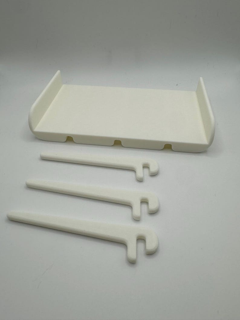 May include: White plastic shelf with three white plastic brackets. The shelf has four holes on the bottom and the brackets have a hook on one end.