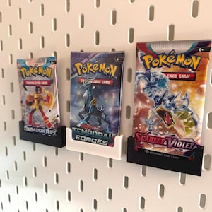 Op de afbeelding: Drie Pok&eacute;mon Trading Card Game booster packs uitgestald op een wit pegboard. De packs worden in zwarte en witte houders gehouden. De packs zijn gelabeld met "Paradox Rift", "Temporal Forces" en "Scarlet & Violet".
