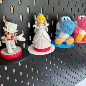 Op de afbeelding: Vier kleine figuurtjes op een zwarte prikbord achtergrond. Een wit gekleed figuurtje van Princess Peach, een blauwe gebreide Yoshi, een roze gebreide Yoshi en een wit gekleed Mario figuurtje.