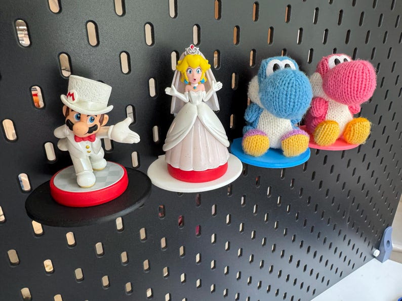 Op de afbeelding: Vier kleine figuurtjes op een zwarte prikbord achtergrond. Een wit gekleed figuurtje van Princess Peach, een blauwe gebreide Yoshi, een roze gebreide Yoshi en een wit gekleed Mario figuurtje.