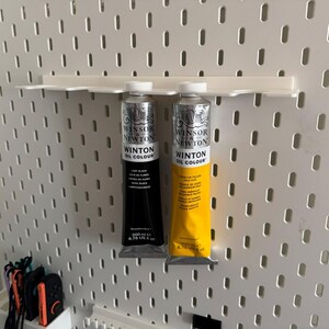 May include: Two tubes of Winton Oil Colour paint, one black and one yellow, displayed on a white pegboard. The black tube is labeled "Lamp Black" and the yellow tube is labeled "Lemon Yellow". Each tube is 200 ml or 6.76 US fl oz.
