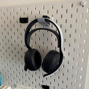 K&ouml;nnte beinhalten: Schwarze und wei&szlig;e Over-Ear-Kopfh&ouml;rer h&auml;ngen an einer wei&szlig;en Lochwand. Die Kopfh&ouml;rer werden von einem grauen Halter gehalten. Die Lochwand hat gleichm&auml;&szlig;ig verteilte L&ouml;cher und ist an einer wei&szlig;en Wand befestigt. Weitere Accessoires sind sichtbar.