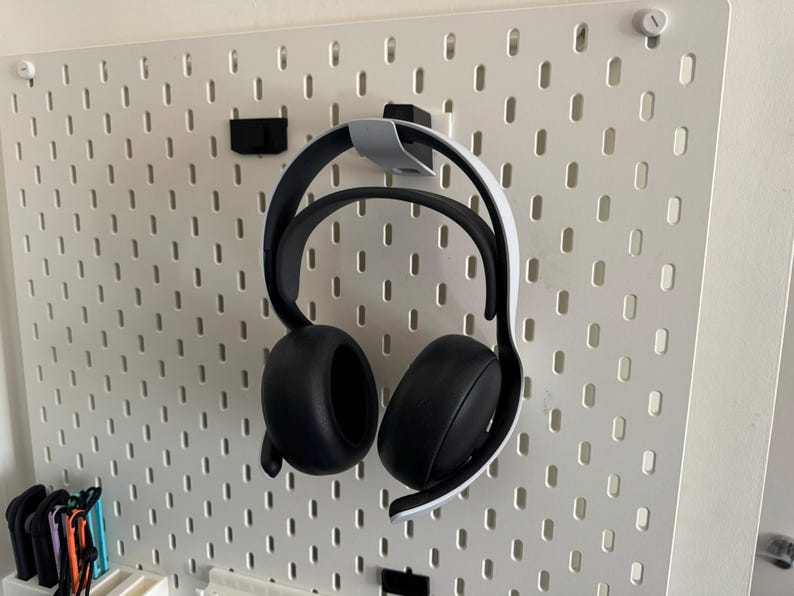 K&ouml;nnte beinhalten: Schwarze und wei&szlig;e Over-Ear-Kopfh&ouml;rer h&auml;ngen an einer wei&szlig;en Lochwand. Die Kopfh&ouml;rer werden von einem grauen Halter gehalten. Die Lochwand hat gleichm&auml;&szlig;ig verteilte L&ouml;cher und ist an einer wei&szlig;en Wand befestigt. Weitere Accessoires sind sichtbar.