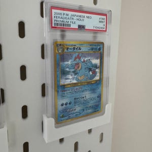 Peut inclure: Une carte Pok&eacute;mon class&eacute;e dans un &eacute;tui de protection transparent. La carte pr&eacute;sente un design bleu et jaune avec du texte japonais et une repr&eacute;sentation d'une cr&eacute;ature ressemblant &agrave; un crocodile. L'&eacute;tui porte une &eacute;tiquette rouge avec le texte "2000 P.M. JAPANESE NEO FERALIGATR-HOLO".