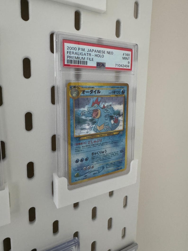 Peut inclure: Une carte Pok&eacute;mon class&eacute;e dans un &eacute;tui de protection transparent. La carte pr&eacute;sente un design bleu et jaune avec du texte japonais et une repr&eacute;sentation d'une cr&eacute;ature ressemblant &agrave; un crocodile. L'&eacute;tui porte une &eacute;tiquette rouge avec le texte "2000 P.M. JAPANESE NEO FERALIGATR-HOLO".