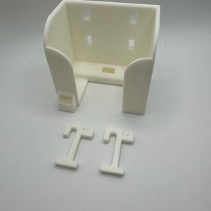 May include: A cream-colored, 3D-printed wall-mounted holder with a rectangular shape and rounded edges. The holder has four rectangular slots and two T-shaped mounting pieces. The design is simple and functional, suitable for various organizational purposes.