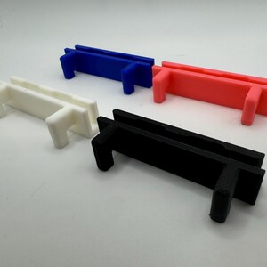 Pode incluir: Quatro suportes de pl&aacute;stico impressos em 3D em branco, preto, azul e rosa. Os suportes s&atilde;o retangulares com um pequeno l&aacute;bio de cada lado e uma pequena aba retangular em cada extremidade.