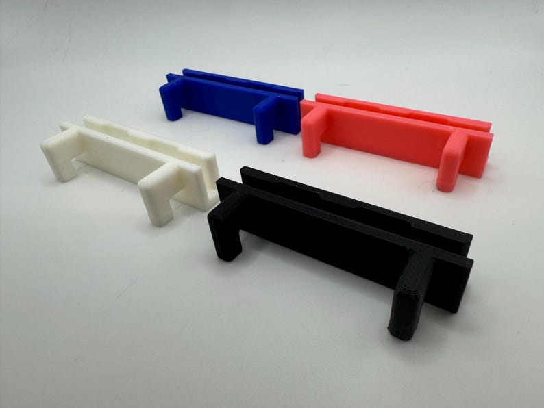 Pode incluir: Quatro suportes de pl&aacute;stico impressos em 3D em branco, preto, azul e rosa. Os suportes s&atilde;o retangulares com um pequeno l&aacute;bio de cada lado e uma pequena aba retangular em cada extremidade.
