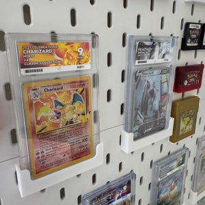 Może przedstawiać: Biała ściana z tablicą perforowaną, na kt&oacute;rej znajdują się liczne ocenione karty Pokemon w ochronnych etui. Karty obejmują Charizarda, Mewtwo i Jolteona. Karty są ocenione na 9 z 10 i są wystawione w przezroczystych plastikowych etui.
