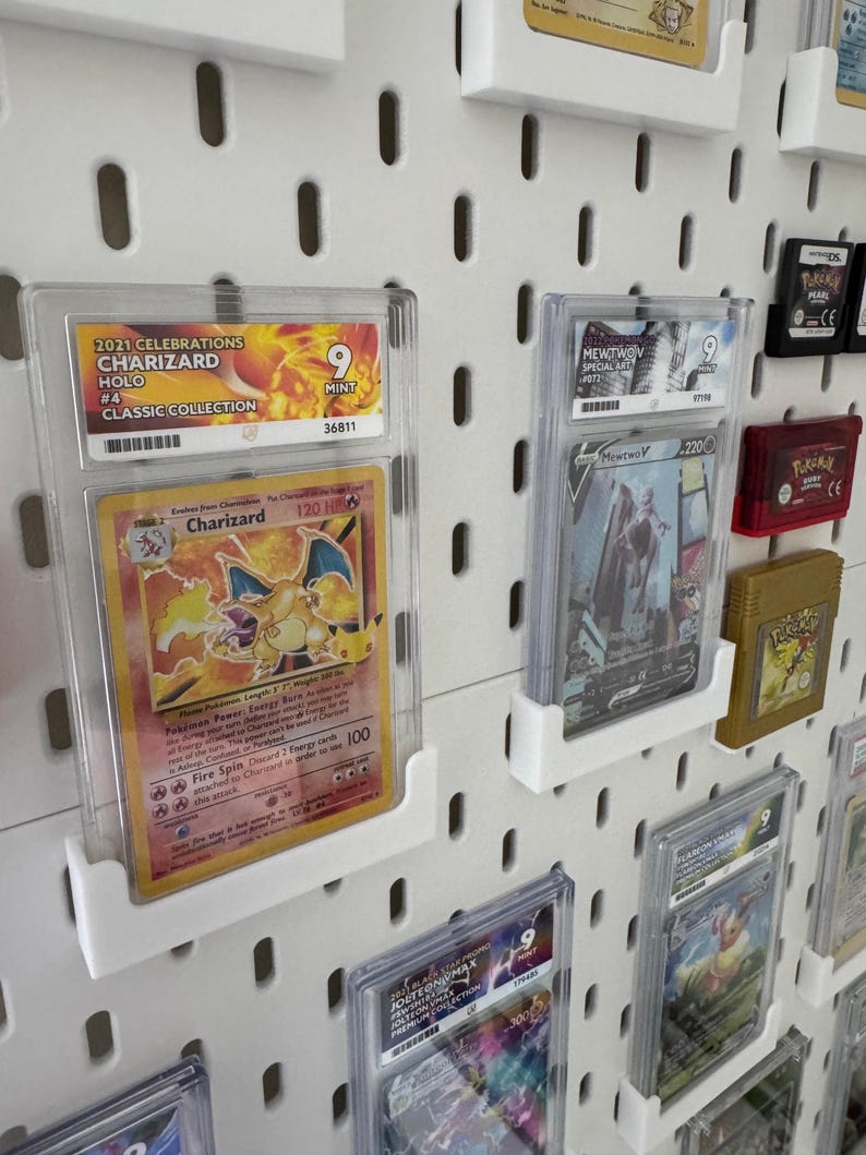 Może przedstawiać: Biała ściana z tablicą perforowaną, na kt&oacute;rej znajdują się liczne ocenione karty Pokemon w ochronnych etui. Karty obejmują Charizarda, Mewtwo i Jolteona. Karty są ocenione na 9 z 10 i są wystawione w przezroczystych plastikowych etui.