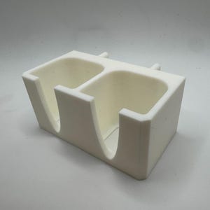 Puede incluir: Un organizador de escritorio rectangular de color crema con dos compartimentos. Cada compartimento tiene una abertura curva en la parte delantera. Dos peque&ntilde;os postes cil&iacute;ndricos est&aacute;n en la parte superior. El organizador es de material mate.