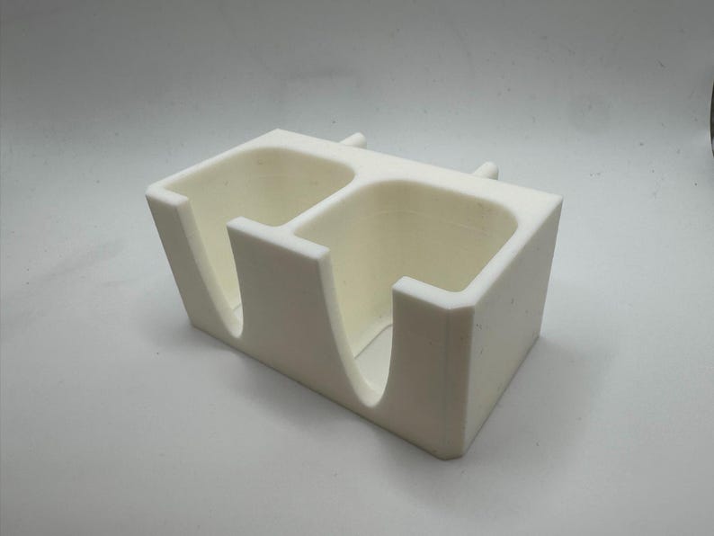 Puede incluir: Un organizador de escritorio rectangular de color crema con dos compartimentos. Cada compartimento tiene una abertura curva en la parte delantera. Dos peque&ntilde;os postes cil&iacute;ndricos est&aacute;n en la parte superior. El organizador es de material mate.