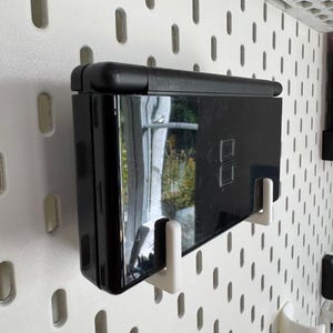 Puede incluir: Una consola de juegos port&aacute;til negra montada en una pared perforada blanca con soportes blancos. La consola est&aacute; cerrada y refleja el entorno. La pared perforada tiene una serie de agujeros ovalados espaciados.