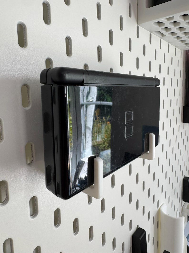Puede incluir: Una consola de juegos port&aacute;til negra montada en una pared perforada blanca con soportes blancos. La consola est&aacute; cerrada y refleja el entorno. La pared perforada tiene una serie de agujeros ovalados espaciados.
