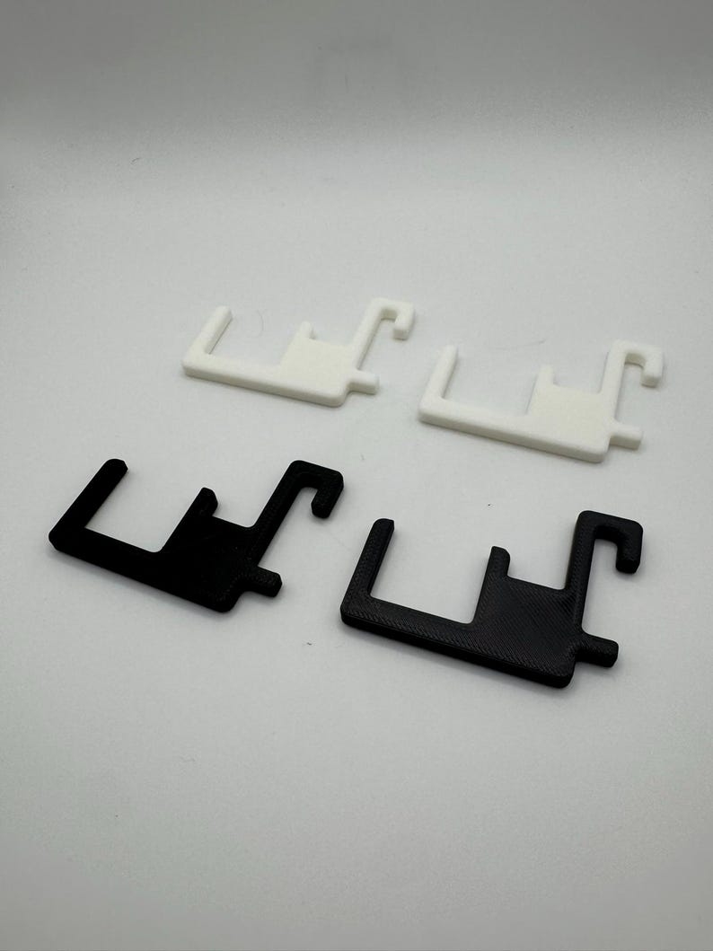 Puede incluir: Cuatro objetos de pl&aacute;stico en forma de soporte. Dos son blancos y dos son negros. Cada uno tiene una base rectangular con una extensi&oacute;n en forma de gancho en un lado y una extensi&oacute;n m&aacute;s peque&ntilde;a e inclinada en el otro. Los objetos est&aacute;n dispuestos en pares.