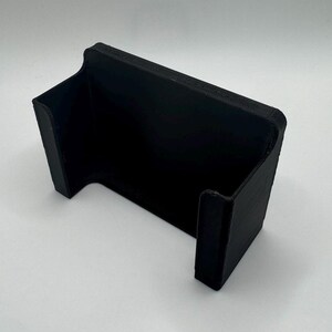 May include: A black plastic phone stand with a curved design. The stand is designed to hold a phone upright.