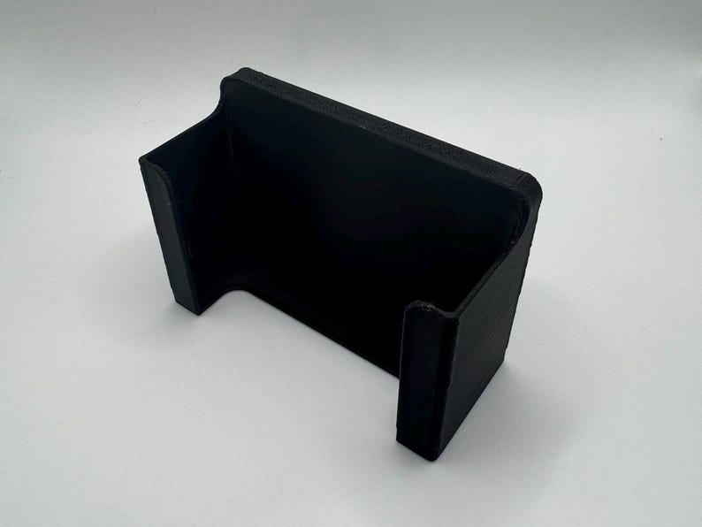 May include: A black plastic phone stand with a curved design. The stand is designed to hold a phone upright.